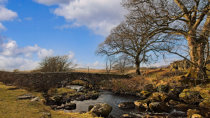 A stream flows over rocky terrain through a winter landscape in Wales, with bare trees silhouetted against blue skies and white clouds. Stone ruins from the historic Ynys y Pandy slate mill are visible, surrounded by moorland and distant hills.