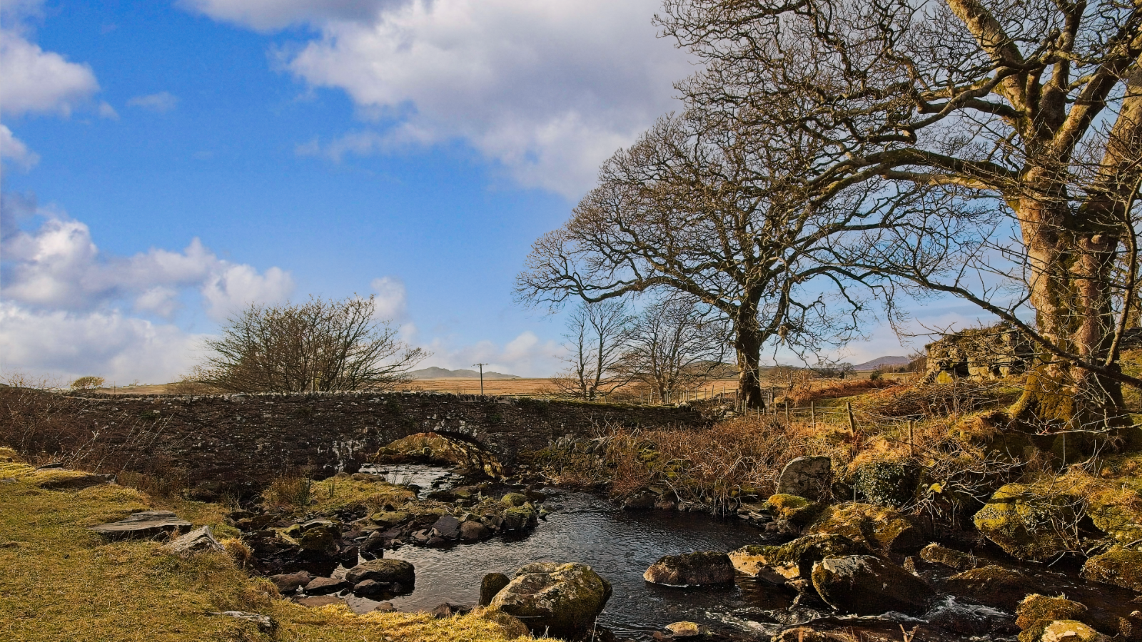 A stream flows over rocky terrain through a winter landscape in Wales, with bare trees silhouetted against blue skies and white clouds. Stone ruins from the historic Ynys y Pandy slate mill are visible, surrounded by moorland and distant hills.