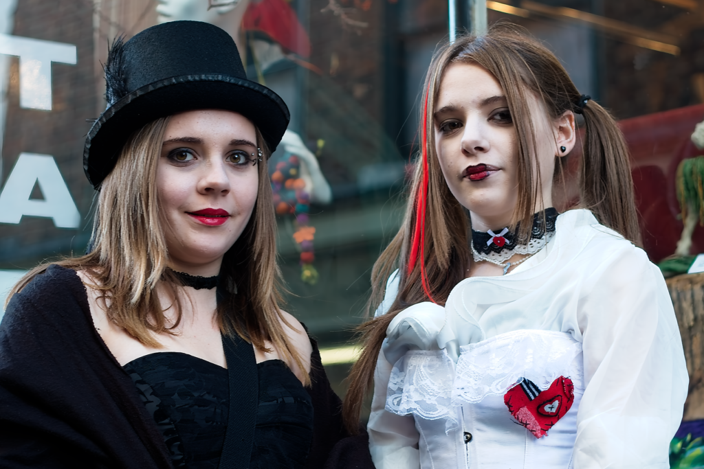 Two young women in alternative gothic fashion pose together - one wearing all black with a top hat and choker, the other in a white ruffled shirt with black bow tie. Both have dramatic makeup with dark eyes and red lips.