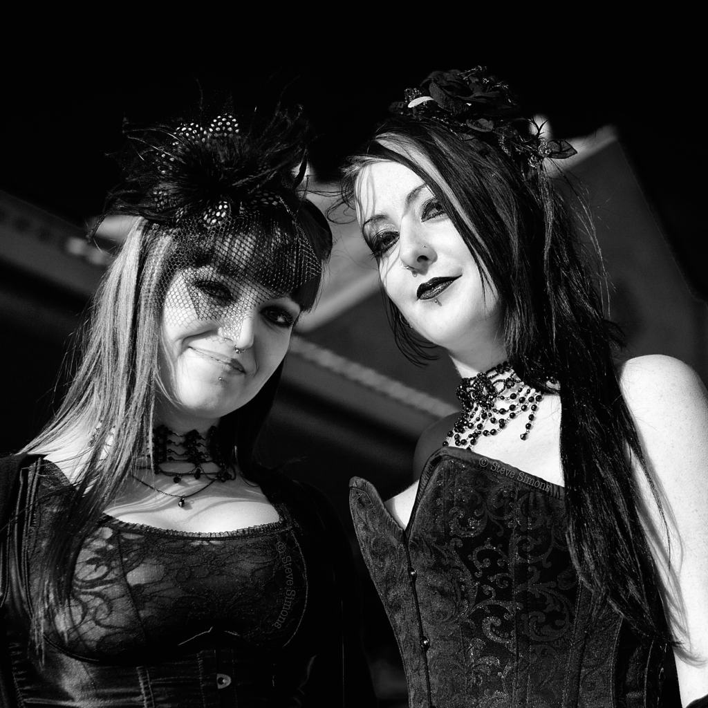 Black and white portrait of two women in elaborate gothic fashion at Whitby Goth Festival. One wears a feathered hat with black netting over her face, the other has long dark hair and an ornate beaded choker with a fitted corset.