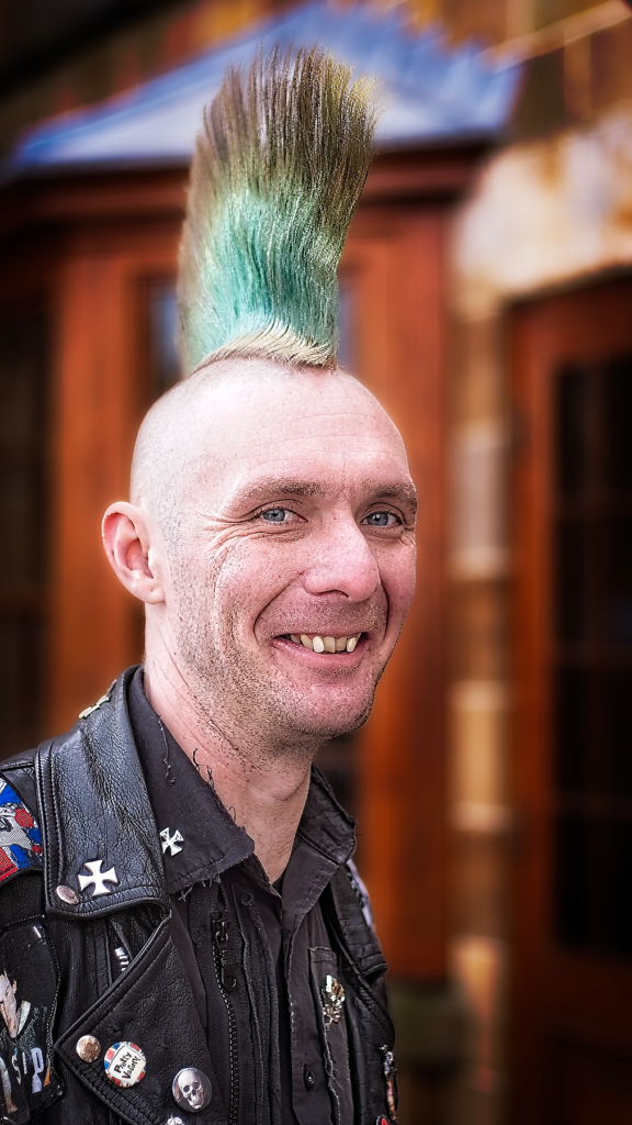 A smiling man with a tall teal and green mohawk hairstyle wearing a black leather jacket with various patches and pins, photographed at Whitby Goth Festival with colorful buildings in the background.