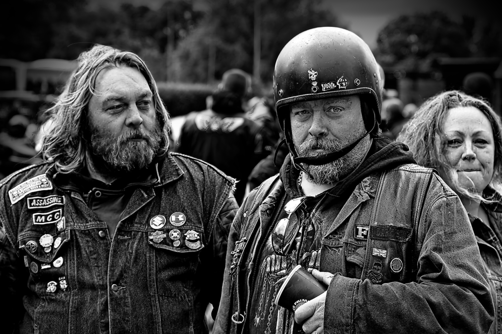 Black and white photograph of two bearded men in leather motorcycle jackets with patches at a protest. One wears a helmet while the other has long hair, both displaying various biker patches and pins on their jackets.