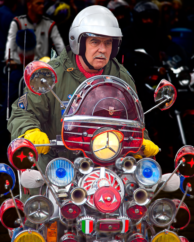 A person in a white helmet sitting on a heavily customized scooter decorated with numerous round mirrors, lights, and chrome accessories. The scooter appears to be a classic Vespa with extensive modifications typical of mod culture.