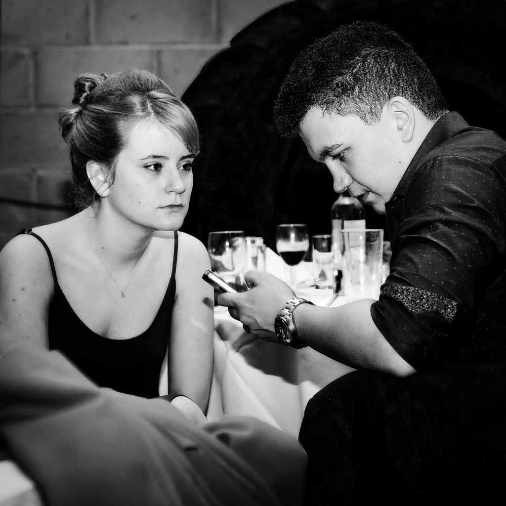 Black and white photograph of a woman in a black dress and a man in dark clothing sitting close together at a table with drinks, captured in an intimate moment of conversation.