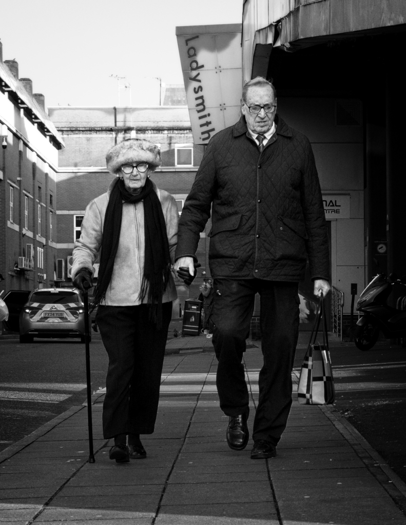 Black and white photograph of an elderly couple holding hands while walking down a street in Ashton-under-Lyne, with shops and buildings visible in the background.