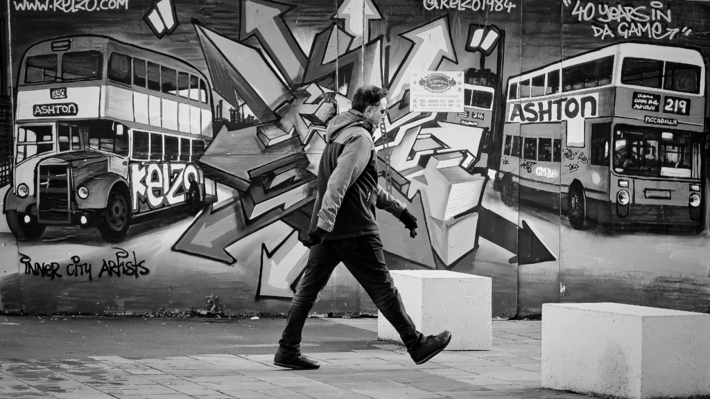 Black and white photograph of a person walking past a wall covered in colorful graffiti and street art, creating a striking contrast between the moving figure and the elaborate artwork.