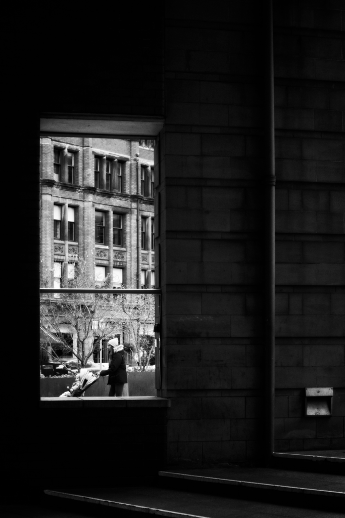 Black and white photograph taken through a modern building's window showing historic Victorian brick buildings across a courtyard on Deansgate in Manchester, with a person walking below.