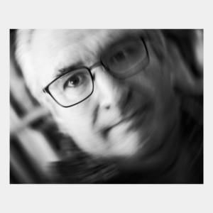 Black and white self-portrait with intentional camera movement creating motion blur and abstract flowing effects, with glasses and facial features still partially recognizable through the artistic blur.