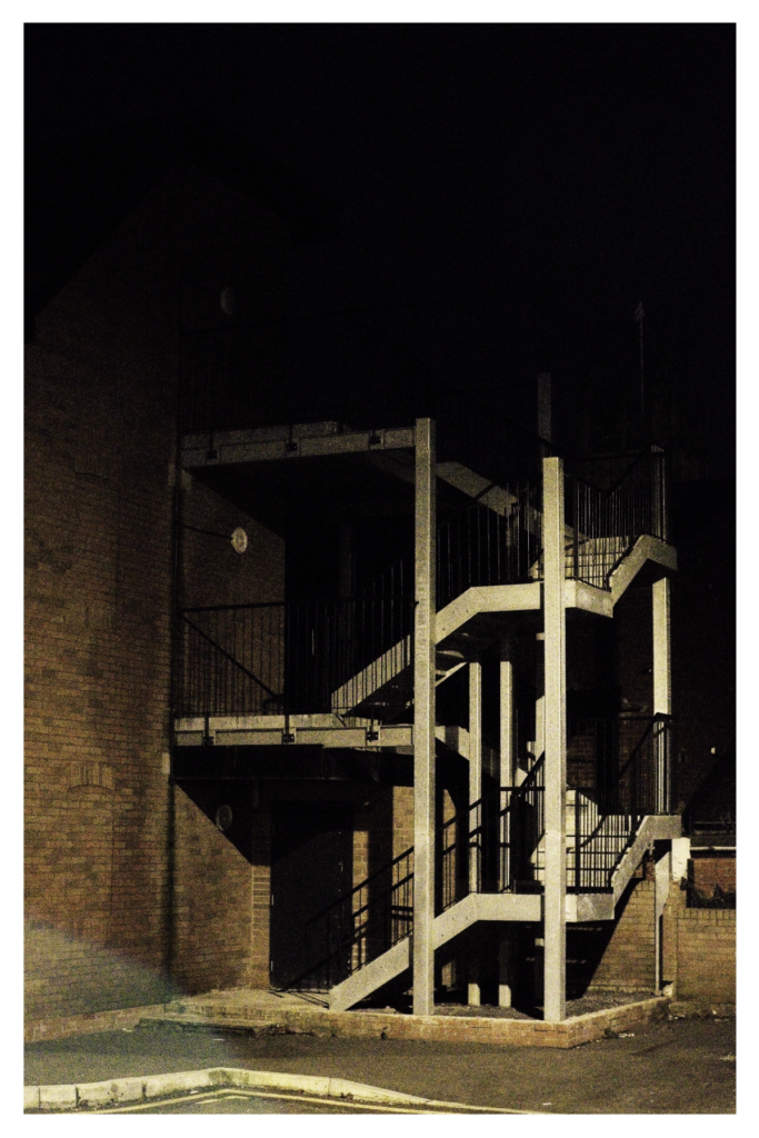 Nighttime photograph of an external metal fire escape staircase with multiple levels, illuminated by artificial lighting against a dark sky in Ashton-under-Lyne.