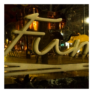 Nighttime photograph showing illuminated metallic letters from the "Fumo" restaurant sign, with reflective surfaces capturing warm city lights on Oxford Road in Manchester.