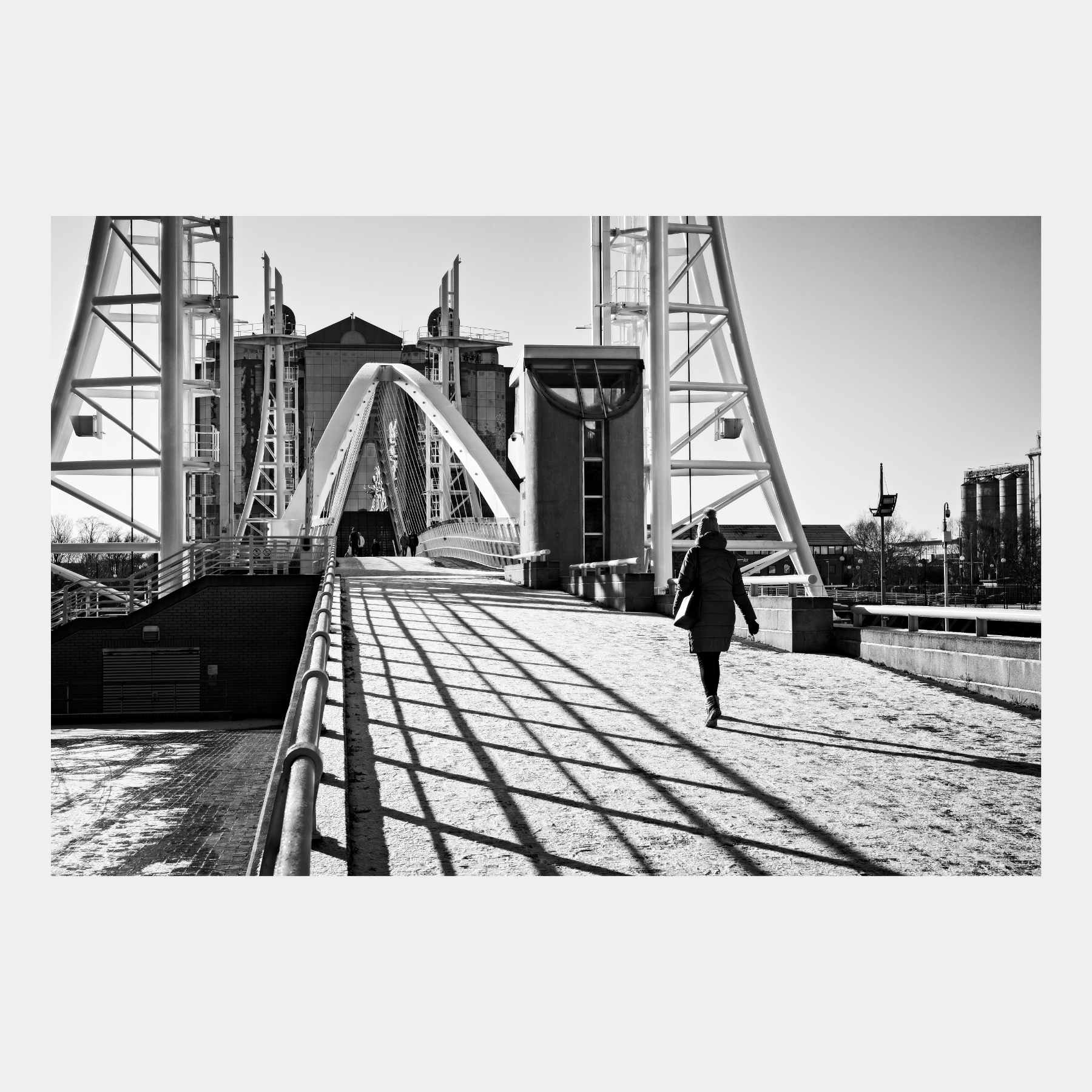 Black and white photograph of a person walking across the Millennium Bridge at Salford Quays, with dramatic shadows from the cable-stay bridge structure creating geometric patterns on the walkway, and MediaCity visible in the background.