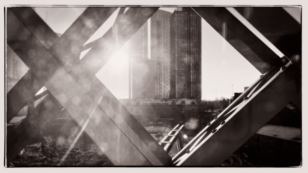 Black and white photograph taken from inside a tram looking up through the geometric ironwork of a bridge at Deansgate, with tower blocks visible through the angular metal framework creating an abstract composition.