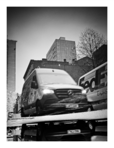 Black and white photograph showing an inverted reflection of a delivery van and city buildings in a rain puddle, creating a surreal upside-down perspective of the urban street scene.