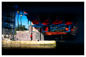 Abstract water reflection photograph showing distorted buildings and urban elements in vivid blues and reds, with rippling water creating an impressionistic effect and a small red-clad figure visible in the composition.