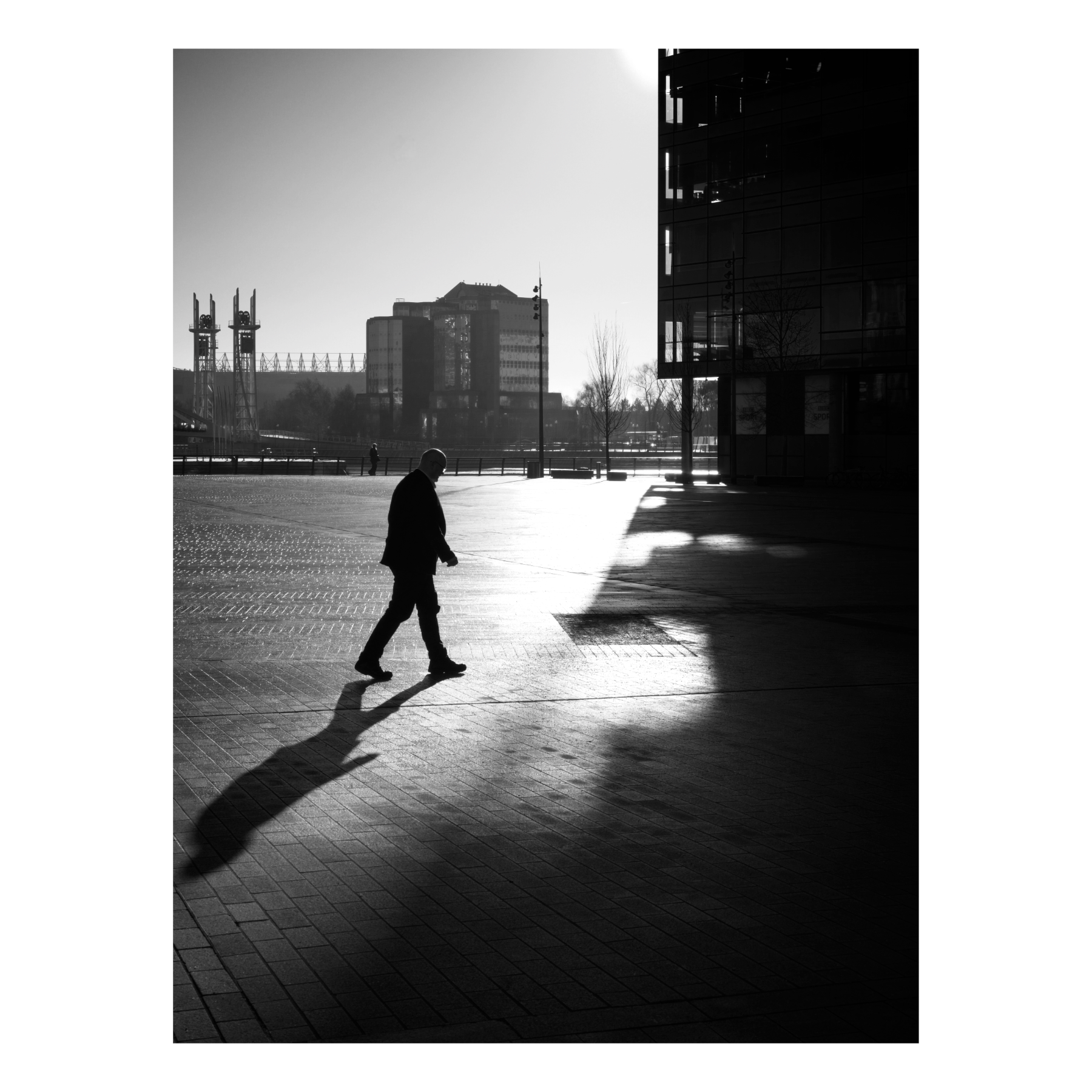 Black and white photograph of a person walking across a plaza at MediaCity in Salford Quays, creating a dramatic silhouette with a long shadow against geometric paved surfaces, with modern buildings in the background.