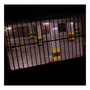 Photograph taken through a metal grated window showing the interior of an underground concrete car park, with warm yellow lighting creating a geometric pattern through the security mesh.