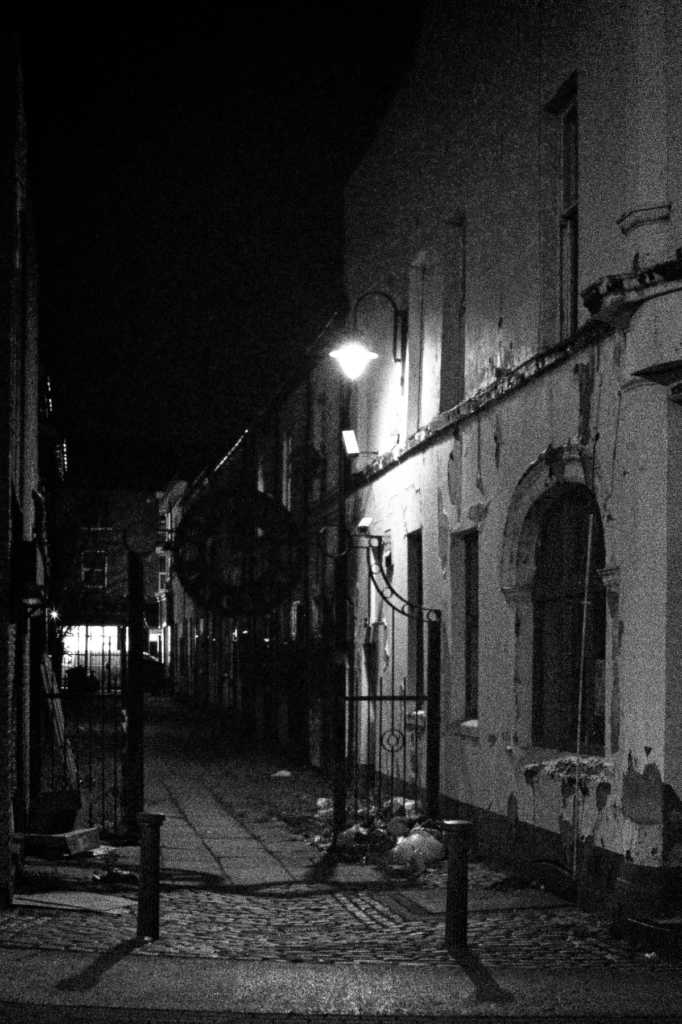 Black and white nighttime photograph of Stamford Arcade in Ashton-under-Lyne showing the covered shopping passage with Victorian ironwork and dramatic lighting creating strong contrasts between light and shadow.