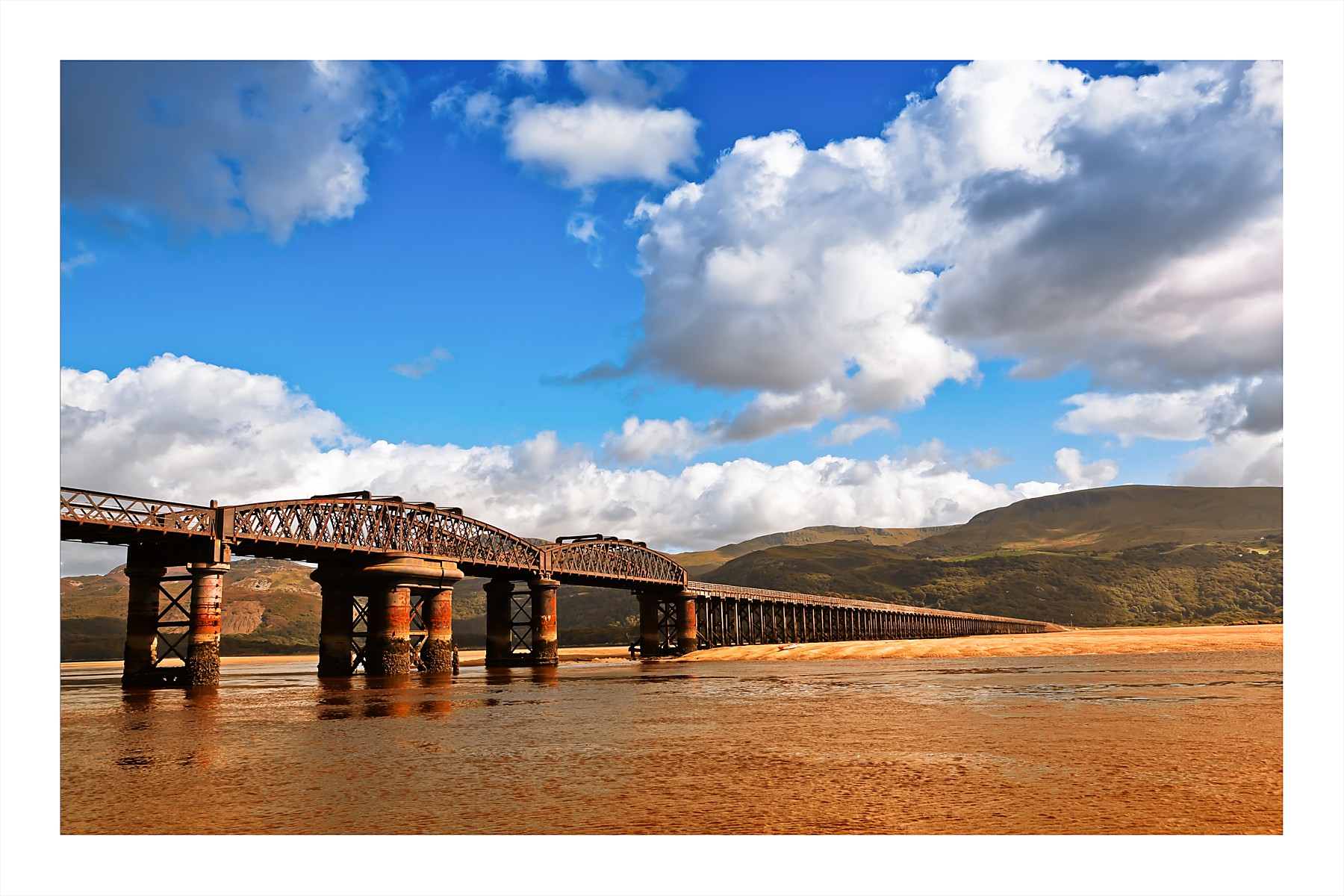 A colour photograph of Barmouth Bridge in Wales, a long Victorian railway viaduct stretching across a tidal estuary. The iron and timber structure recedes into the distance over golden-brown sand and shallow water at low tide. Green hills rise behind the bridge under a bright blue sky with large white clouds.