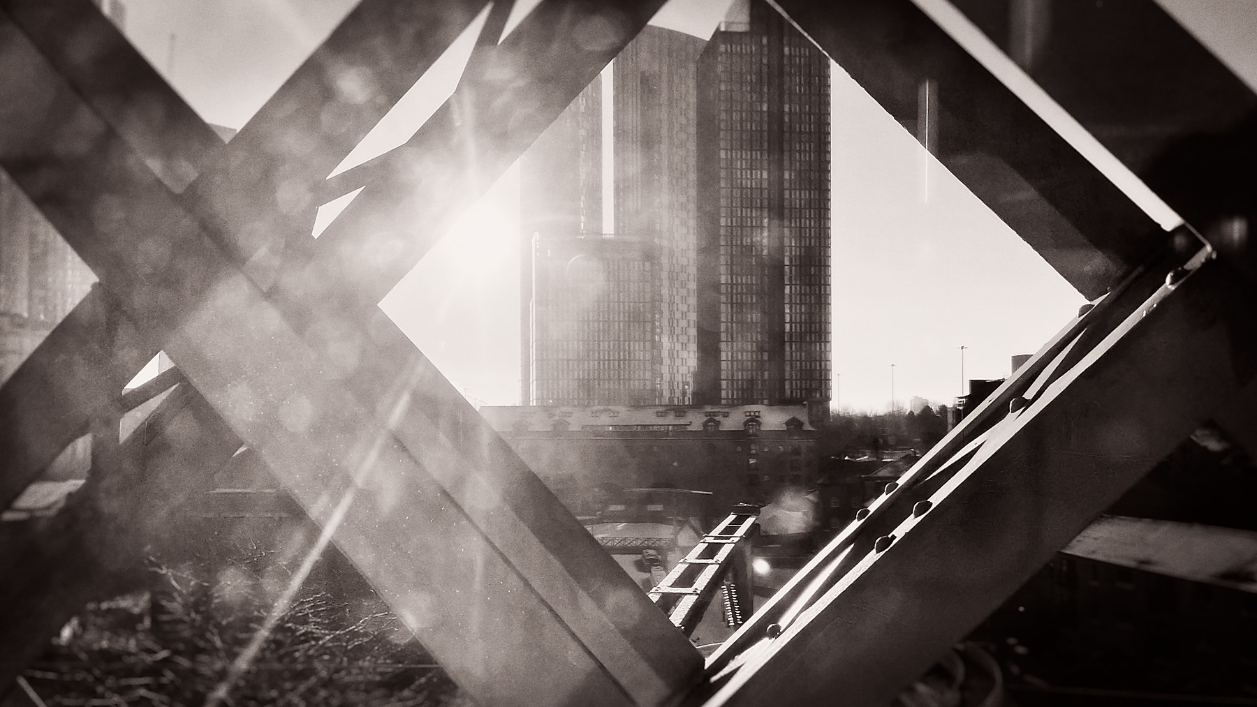 Black and white photograph taken from inside a tram looking up through the geometric ironwork of a bridge at Deansgate, with tower blocks visible through the angular metal framework creating an abstract composition.