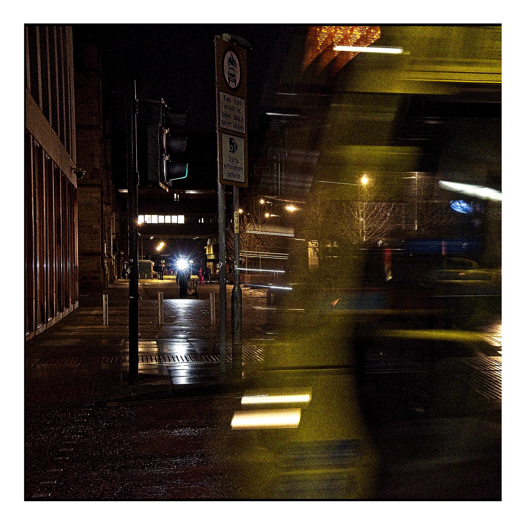 Nighttime street photograph showing a blurred tram or bus in motion through Manchester city centre, with light trails from the moving vehicle and wet pavement reflecting street lights.