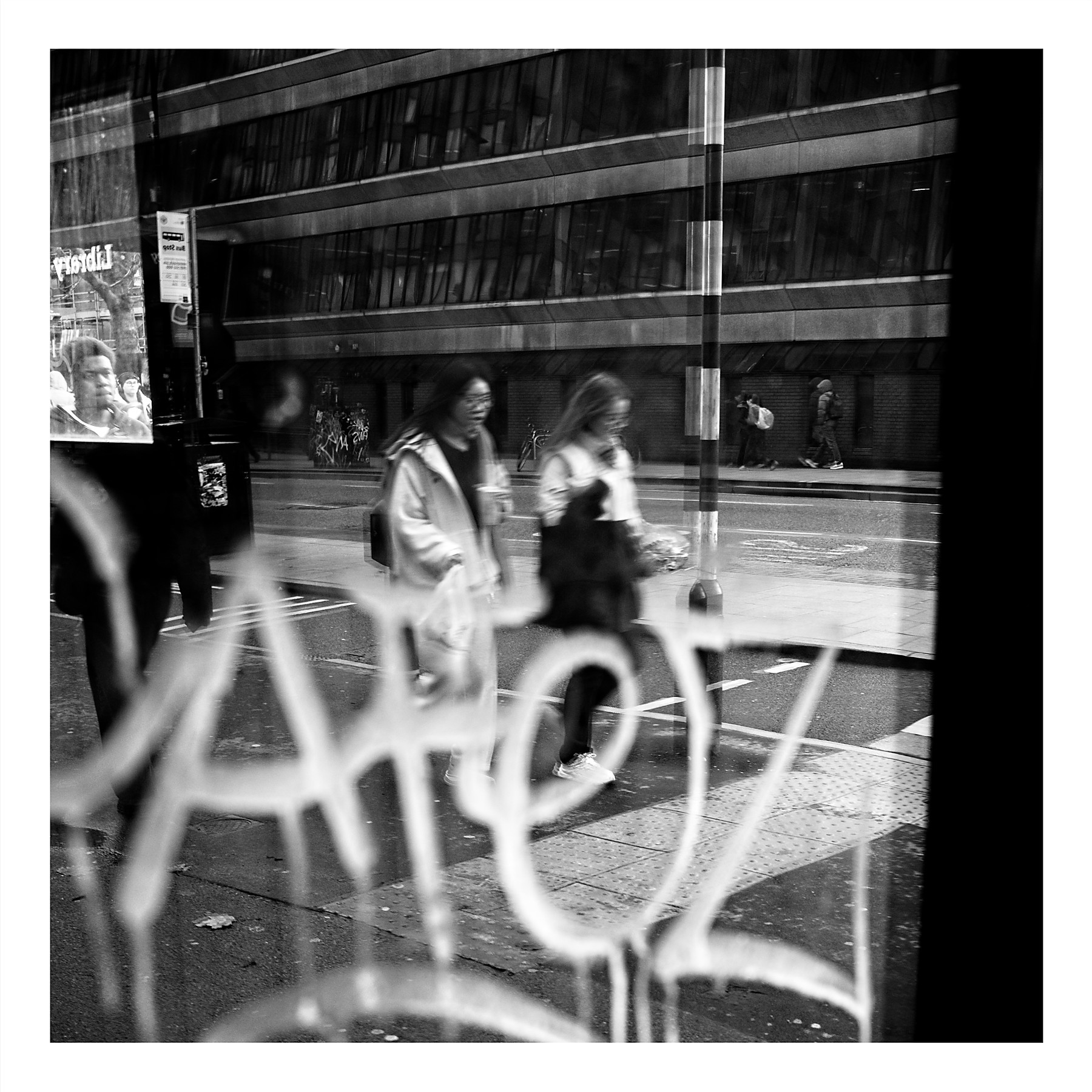 Black and white photograph taken through a shop window showing blurred figures walking in a reflected street scene, with the actual window frame visible in the top left, creating a complex layered composition of reflections and reality.