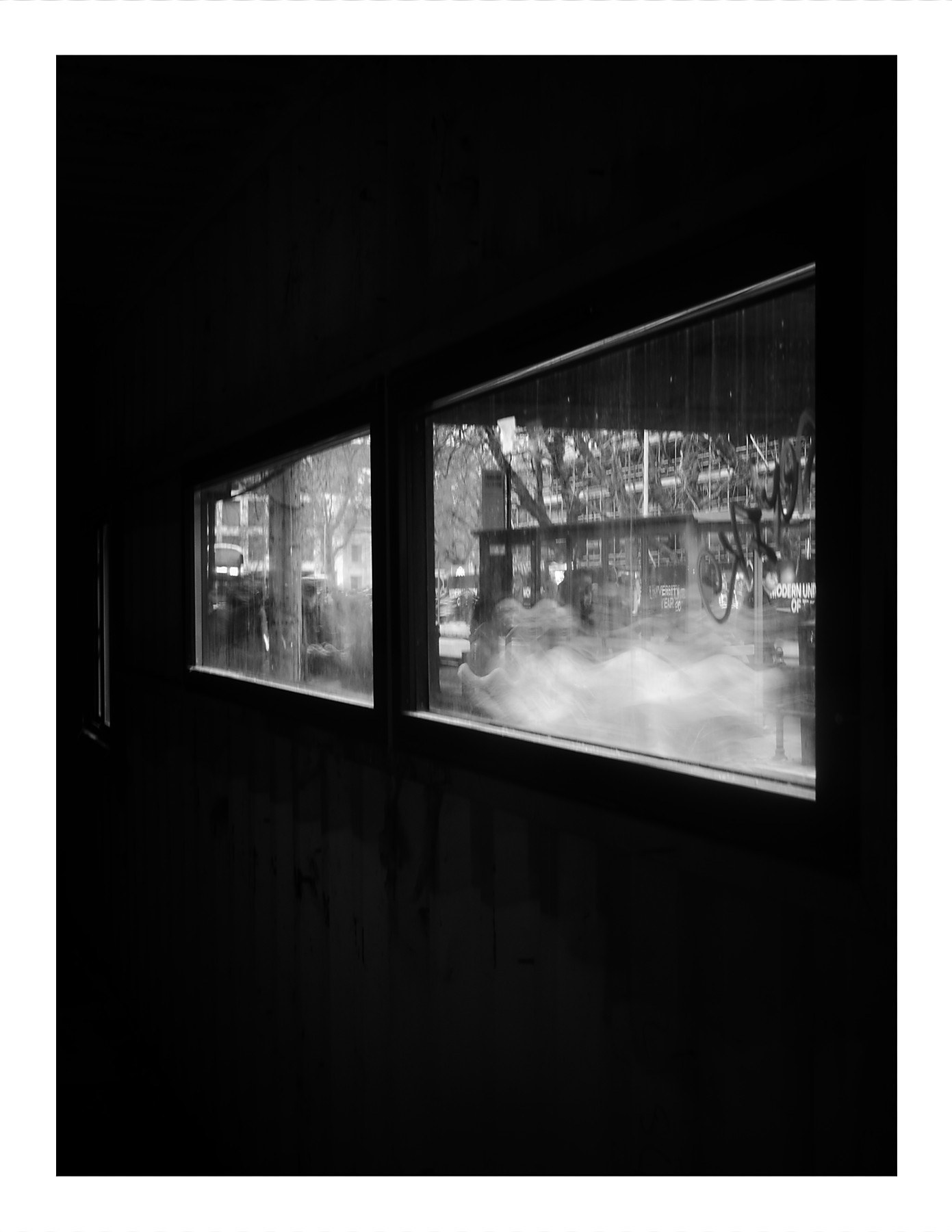 Black and white photograph taken from a dark interior looking through windows at people passing outside, with slow shutter speed creating flowing, blurred motion of the pedestrians.