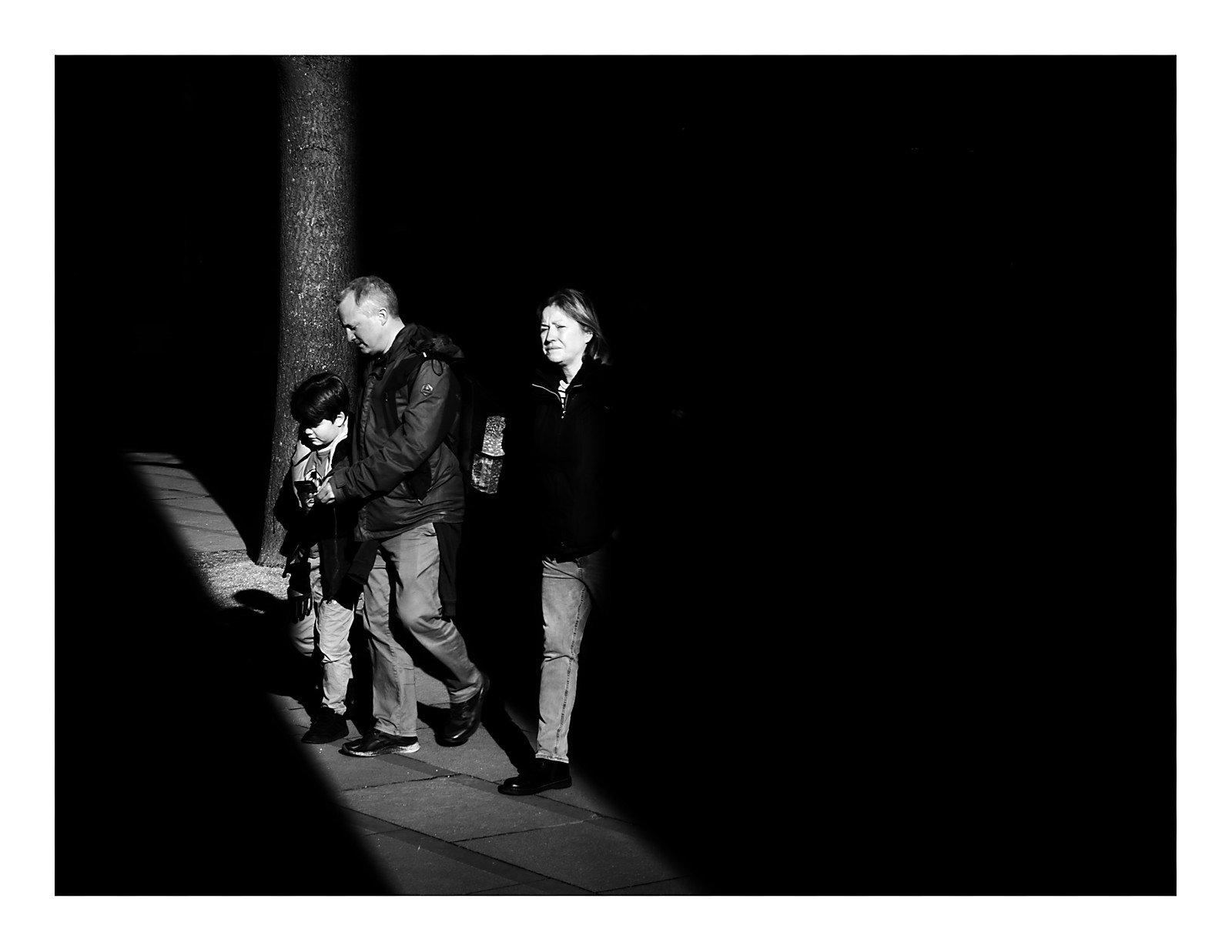 Black and white photograph of a family with young child crossing through a dramatic sunbeam between harsh shadows in St Peter's Square Manchester, creating striking contrast between light and dark areas.