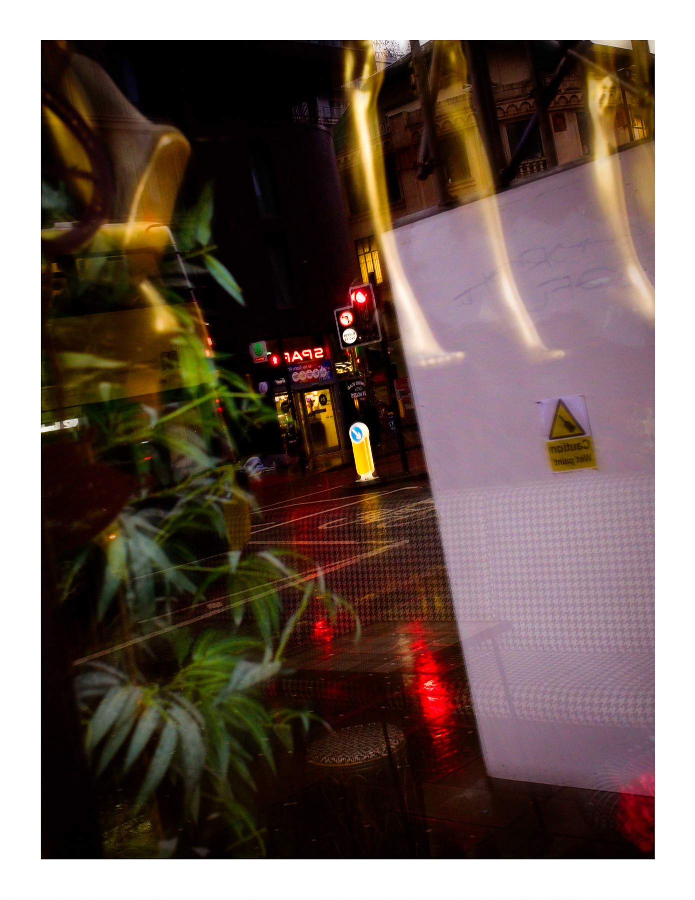 A rain-soaked city street at night, viewed through a glass window with reflections. A tropical plant is reflected in the foreground. In the background, a red traffic light glows with its reflection visible on the wet road, a SPAR store sign is lit up, and a white construction hoarding with a yellow caution sign stands to the right.