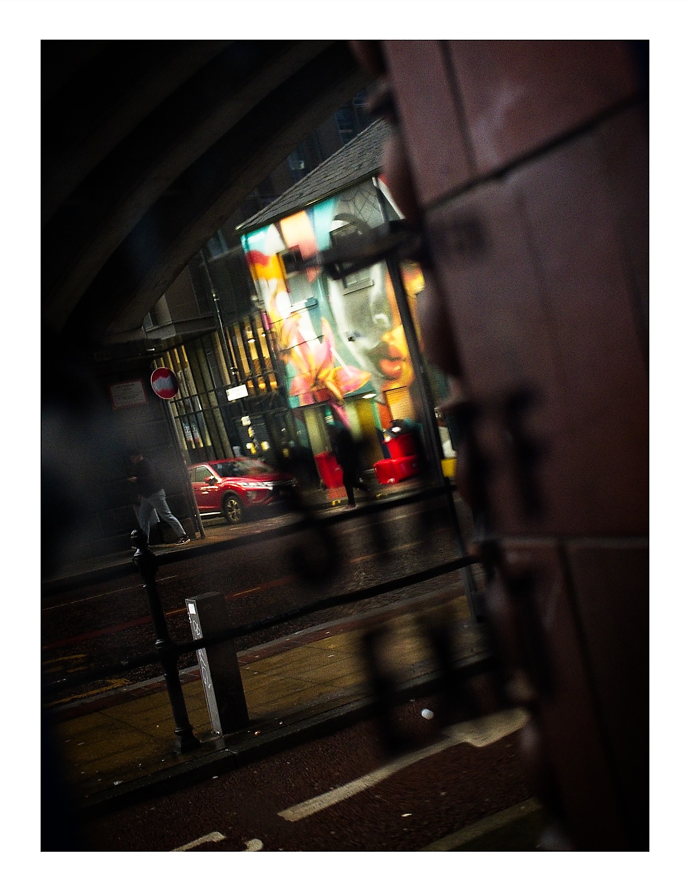A dark, wet city street at night framed by a doorway or gate in the foreground. A colourful mural lights up a building across the street. A pedestrian walks on the pavement beside a parked red car, and a blurred figure stands near the mural. Metal railings line the wet road in the middle distance.