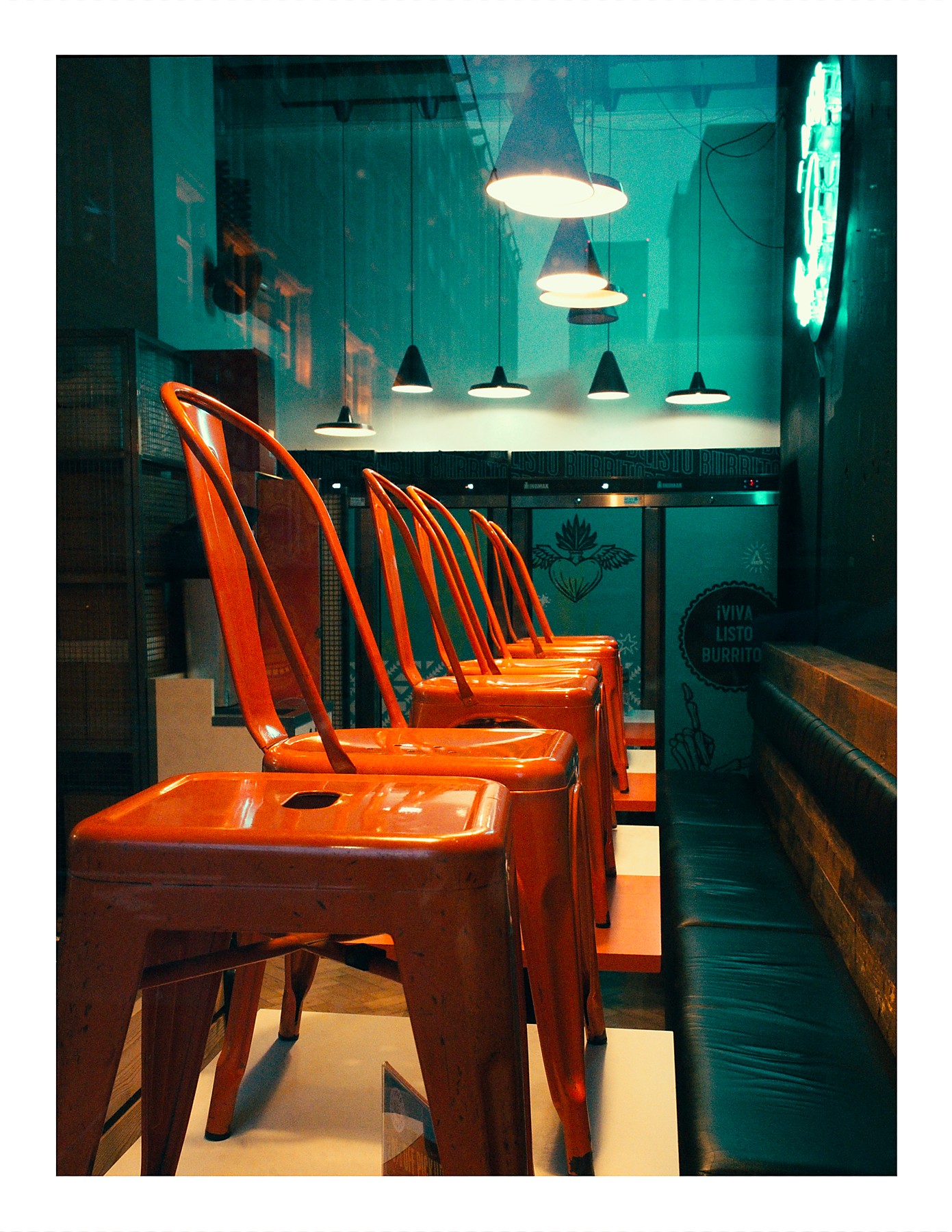 The interior of a burrito restaurant with a row of orange metal chairs along a narrow dining area. Pendant lights hang from a teal-coloured ceiling. A sign on the wall reads "¡Viva Listo Burrito" and a neon sign glows in the upper right. A leather bench seat runs along the right wall.