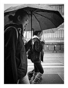 Black and white photograph of students sharing an umbrella while walking in the rain on Oxford Road Manchester, with wet pavement and urban buildings visible in the background.