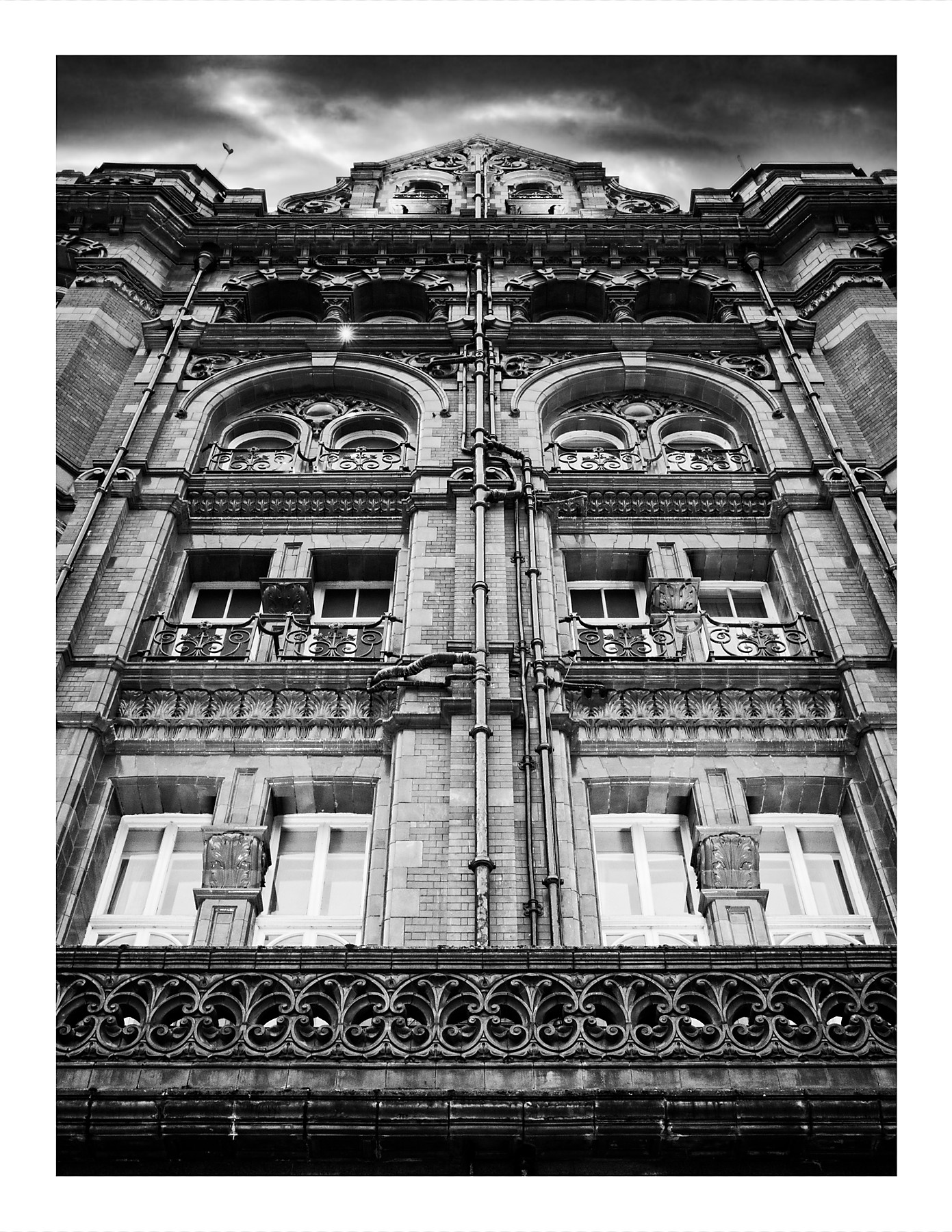 Black and white low-angle photograph of the ornate Victorian façade of the Midland Hotel in Manchester, showing elaborate stonework, decorative balconies, classical arched windows, and intricate architectural details against a dramatic sky.