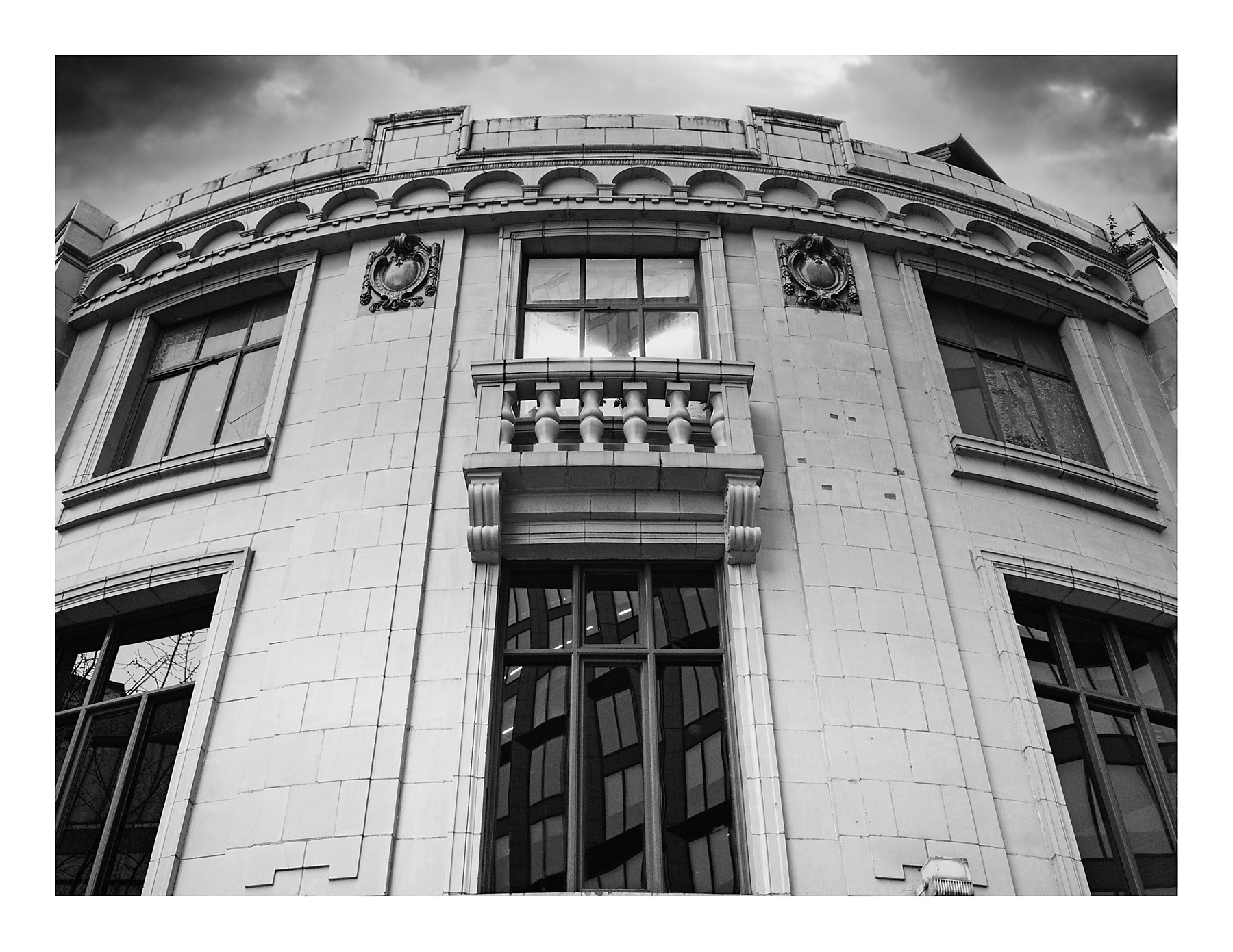 A black and white photograph looking up at the curved corner of the Dancehouse building in Manchester. The pale stone facade features a small balcony with decorative balusters, carved ornamental medallions, arched detailing along the roofline, and large windows reflecting the surrounding buildings. A dramatic cloudy sky fills the background.
