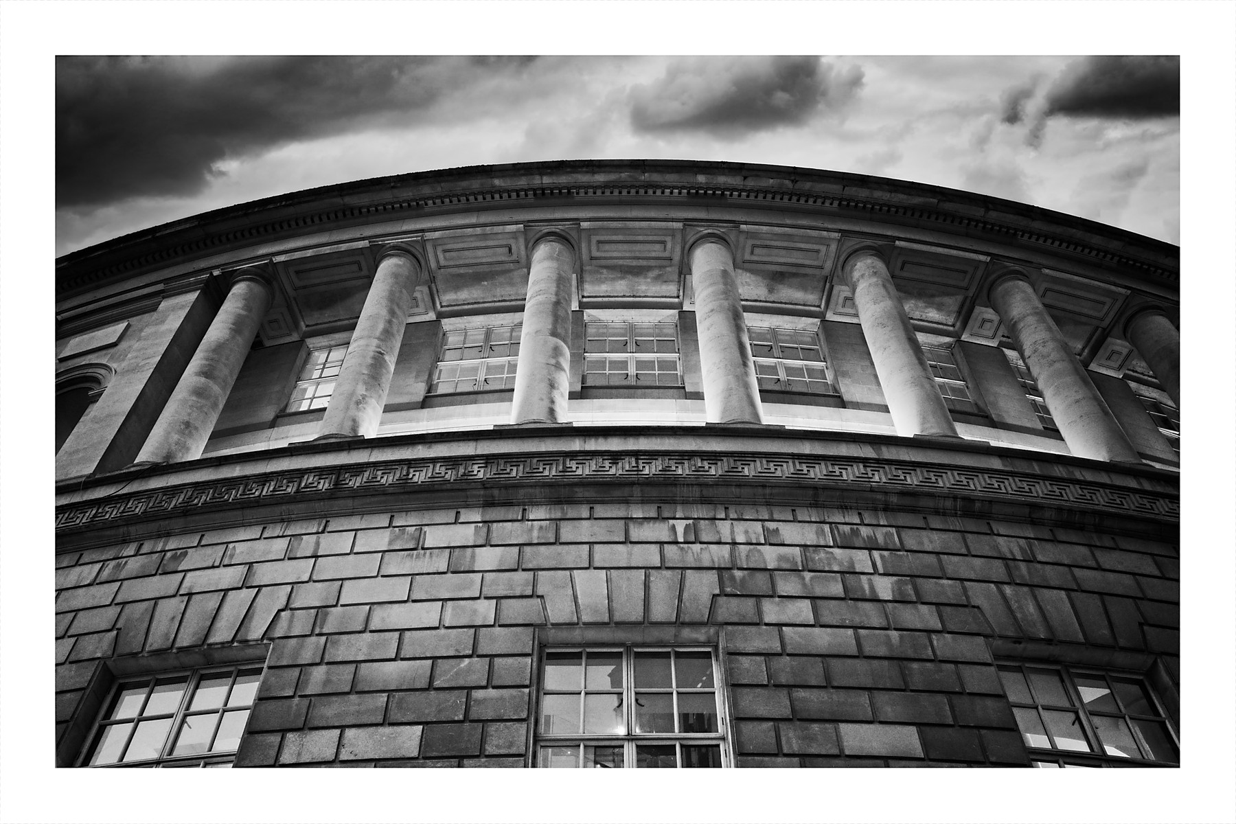 Black and white low-angle photograph of Manchester Central Library showing its curved neoclassical façade with tall Corinthian columns, ornate stonework, and geometric masonry patterns against a dramatic cloudy sky.
