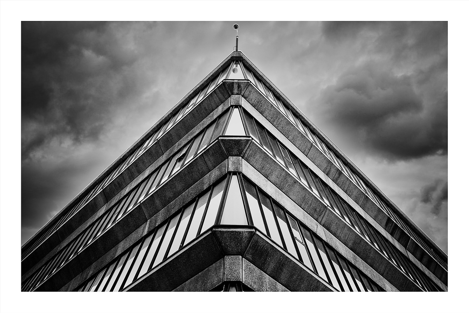 A black and white photograph looking up at the sharp pointed corner of Manchester Metropolitan University's library building. The brutalist facade features strong repeating horizontal lines of concrete and glass that converge symmetrically to a point, with a CCTV camera mounted at the apex. A dramatic cloudy sky fills both sides of the frame.