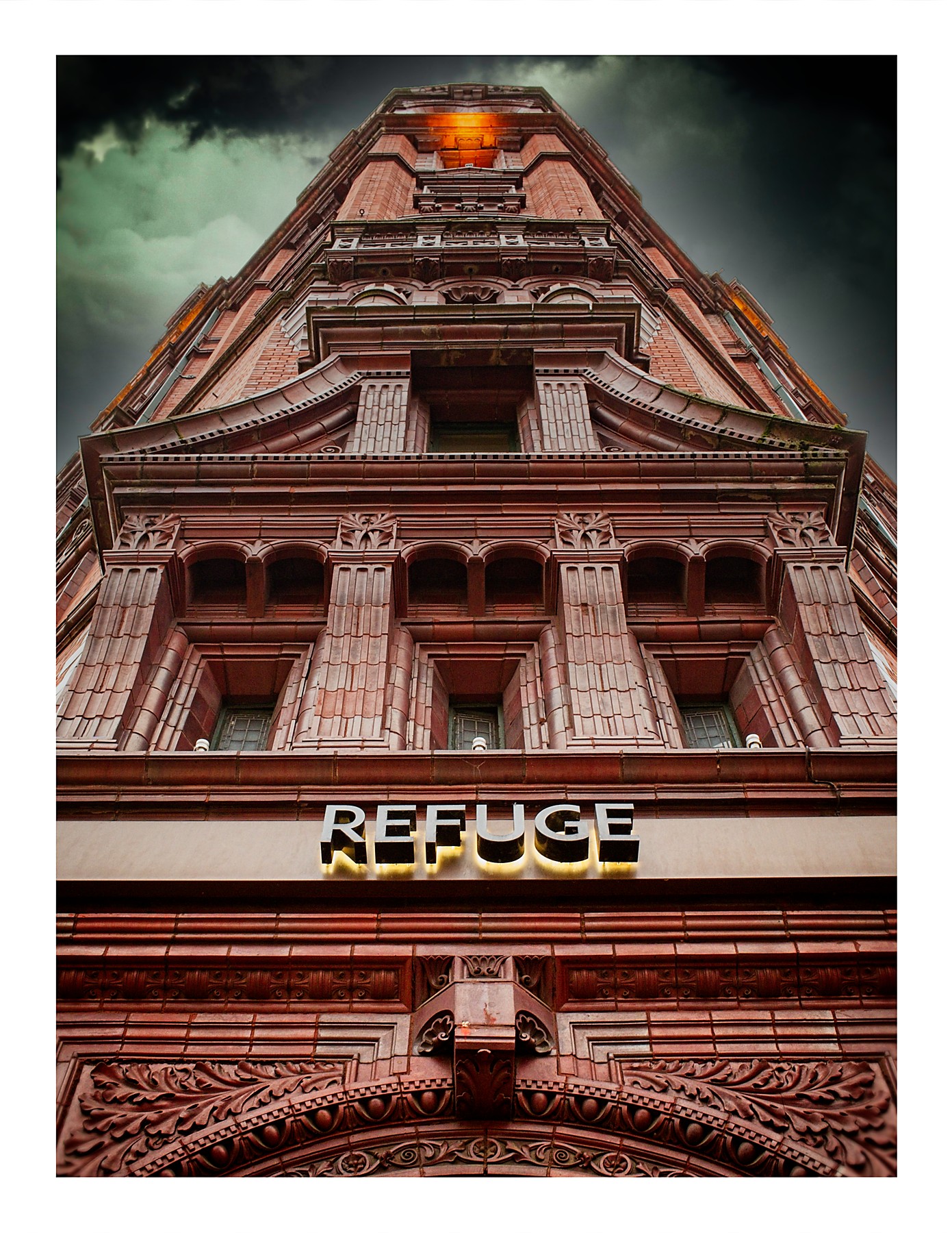 A dramatic upward-looking photograph of the Refuge Building in Manchester. The ornate red terracotta Victorian facade rises steeply towards a glowing lantern at the top of the tower, framed by a dark, moody sky. A illuminated "REFUGE" sign is mounted on the facade in the lower portion of the frame, above richly carved decorative stonework.