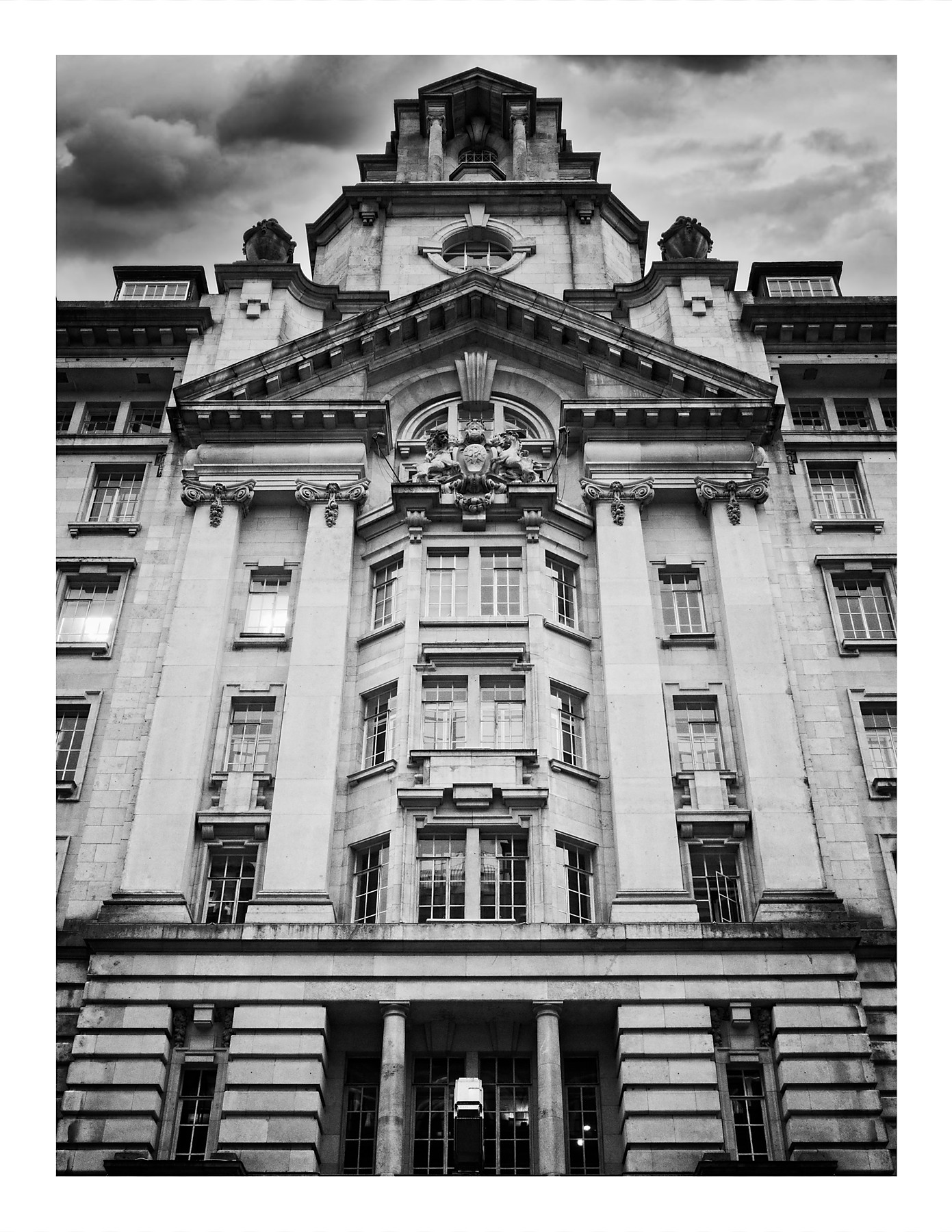 A black and white photograph looking up at the grand Baroque-style facade of St James's Building in Manchester. The ornate stone frontage features large columns, decorative carved stonework including cherubs and scrollwork, arched windows, and a stepped pediment at the top. A dramatic cloudy sky fills the background.