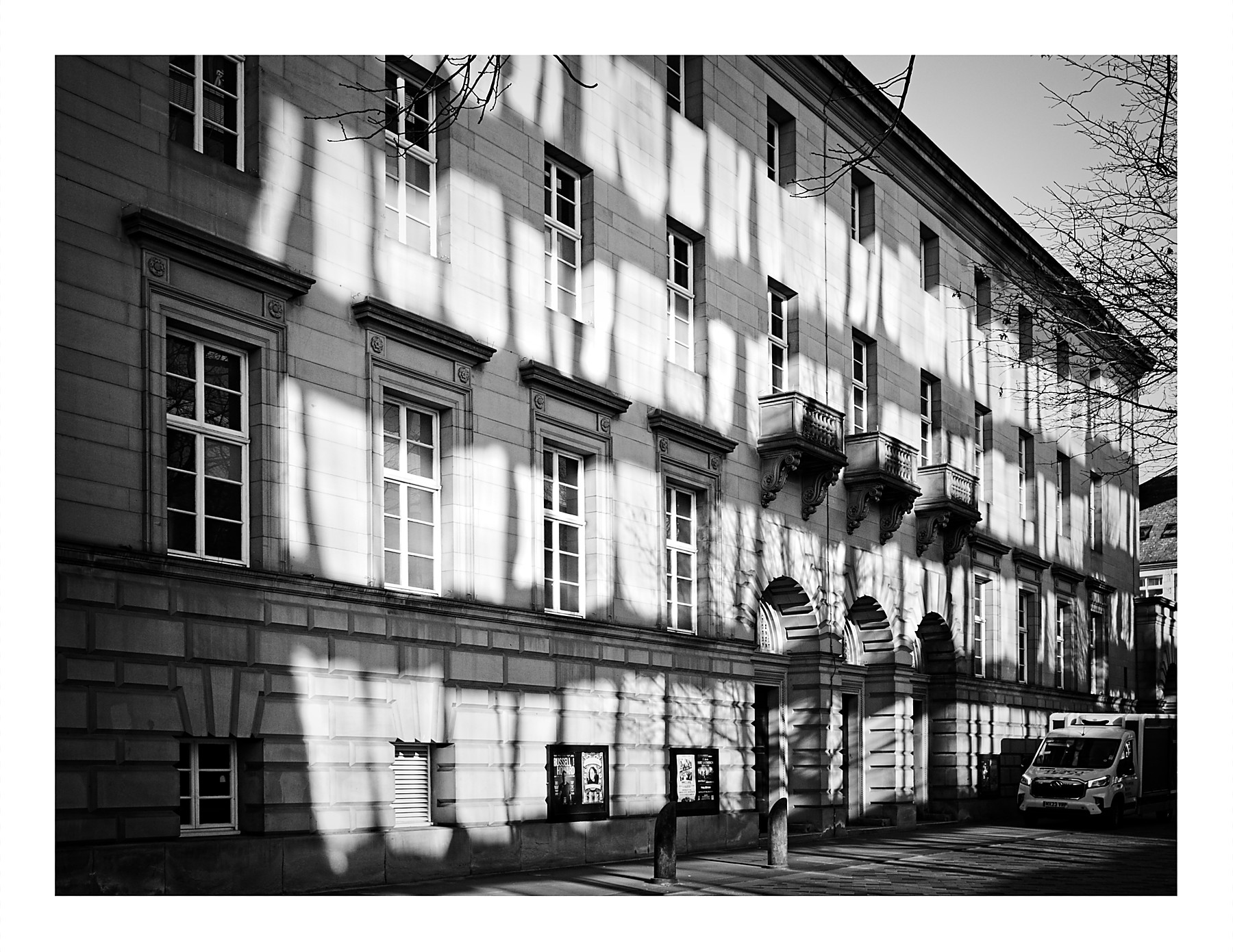 A black and white photograph of the side facade of Sheffield City Hall. Large rectangular patches of reflected sunlight from windows across the street illuminate the pale stone wall, creating bold abstract light patterns across the surface. The classical building features rows of sash windows, ornate carved brackets, and an arched doorway. Bare winter trees cast additional shadows on the wall. A van is parked to the right and event posters are displayed near the entrance.