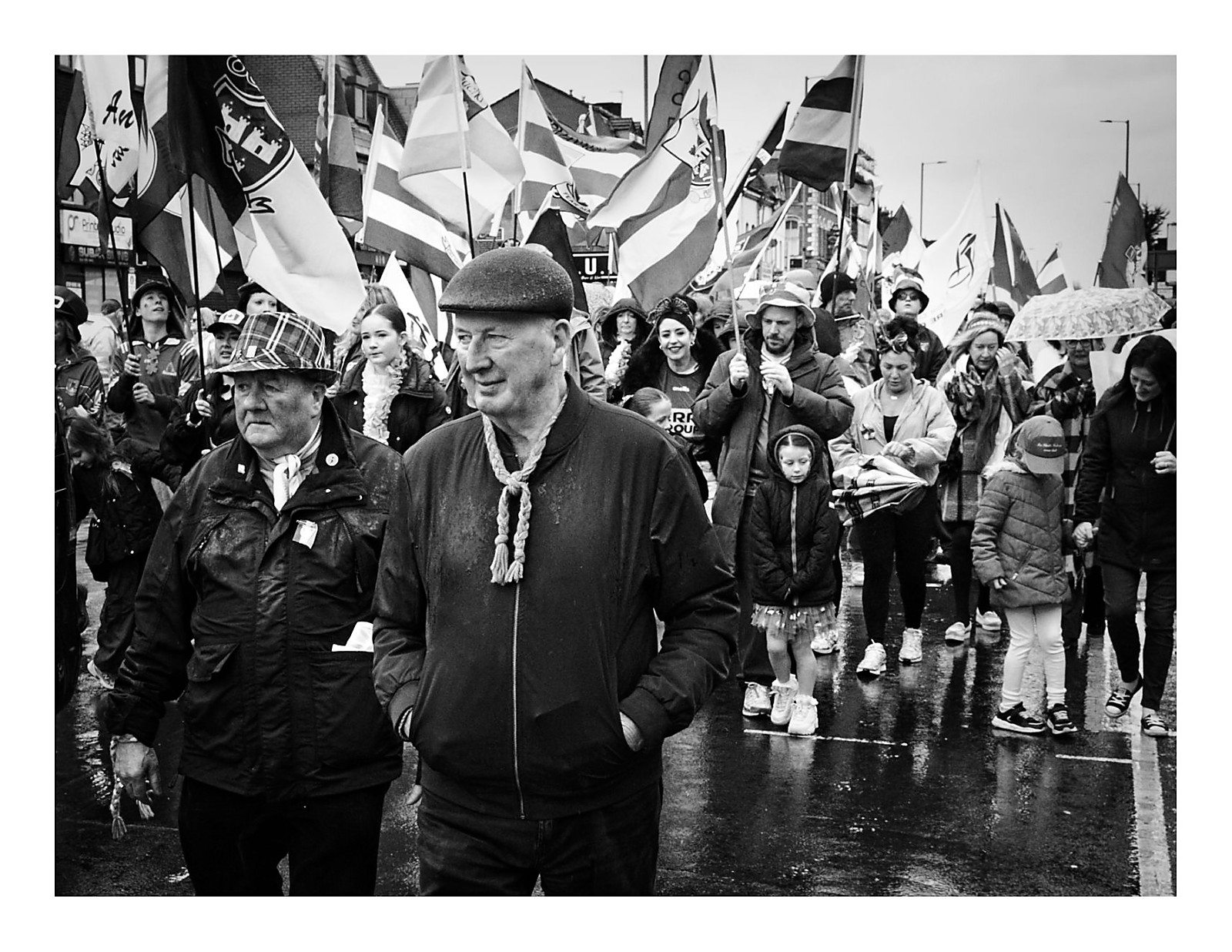 Black and white photograph of two older men leading the community section of the St Patrick's Day parade on Cheetham Hill Road in Manchester in the rain, with a crowd of flag-carrying participants of all ages stretching behind them along the wet road.