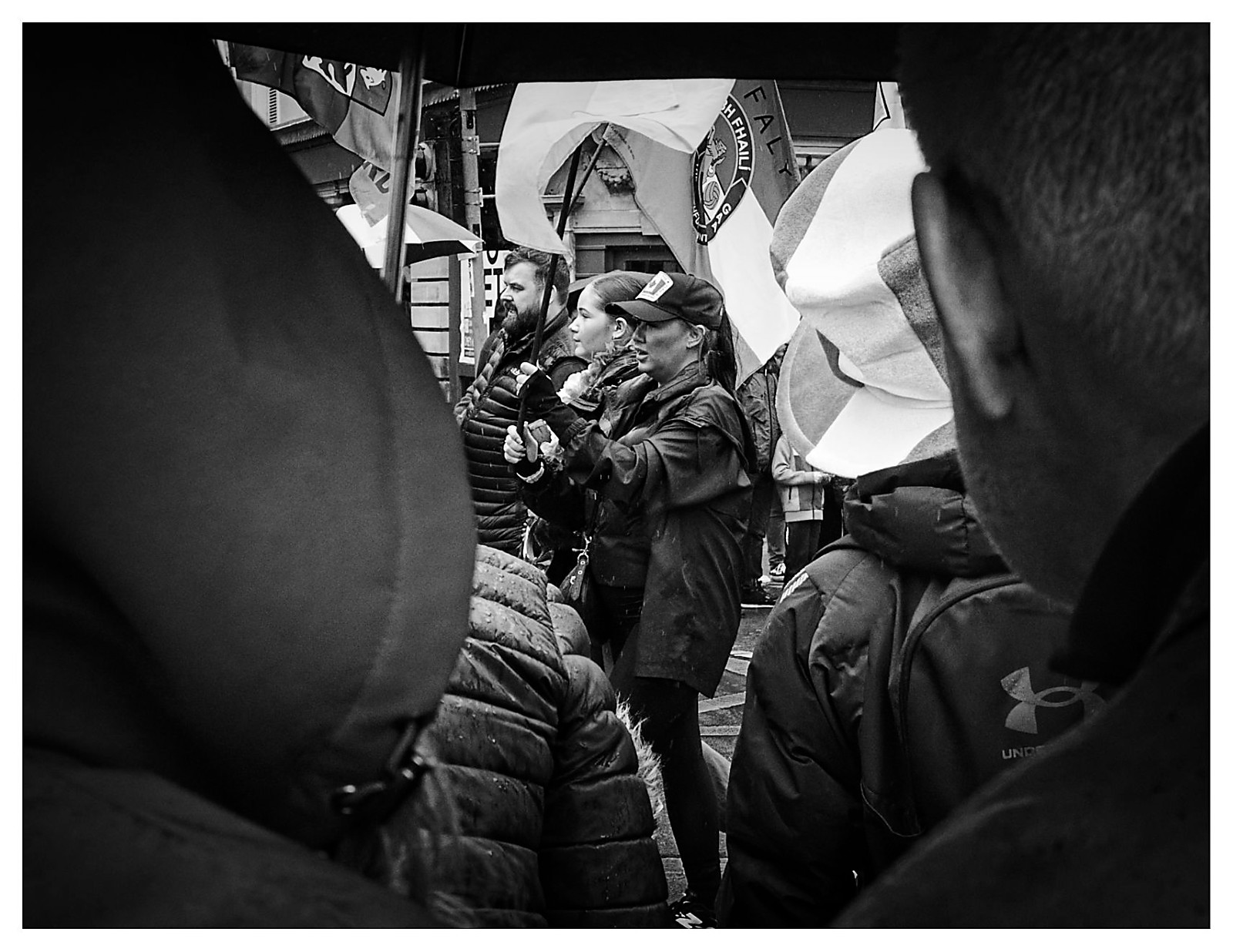 Black and white photograph shot through a gap between two people in a crowd at the St Patrick's Day parade in Manchester, with a young woman in a baseball cap and a bearded man visible in the middle distance, large parade flags billowing above them.