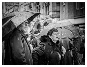 Black and white photograph of a bearded man in glasses and a woman with a polka-dot umbrella standing in the rain at a protest in Manchester city centre, the woman appearing to chant, with a crowd and Victorian stone arches visible behind them.