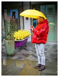 Colour street photograph of a man in a red puffer jacket standing in the rain looking at his phone under a yellow umbrella, beside a purple clawfoot bathtub used as a planter filled with yellow flowers, on a wet city pavement.