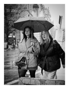 Black and white street photograph of two women sharing an umbrella in heavy rain on Cross Street in Manchester, one in a quilted coat looking at the camera, the other in a puffer jacket grimacing against the weather, with wet pavements and a grand stone building behind them.