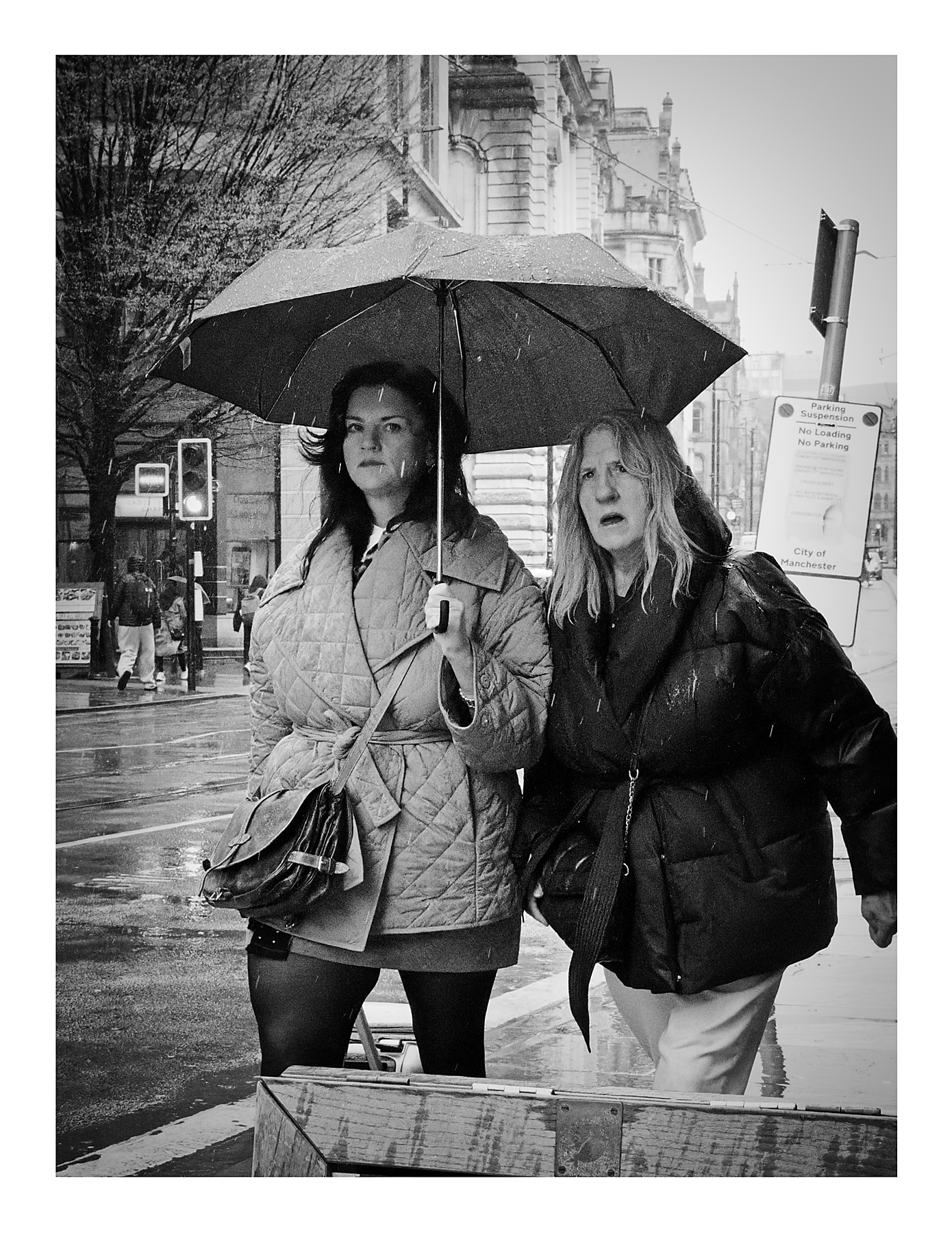 Black and white street photograph of two women sharing an umbrella in heavy rain on Cross Street in Manchester, one in a quilted coat looking at the camera, the other in a puffer jacket grimacing against the weather, with wet pavements and a grand stone building behind them.