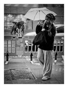Black and white street photograph of a young woman on Corporation Street in Manchester holding a pale umbrella and a large black bag in the rain, wearing a hoodie, wide-leg joggers and a patterned bandana, with crowd control barriers and other pedestrians behind her.