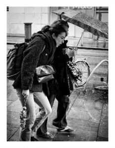 Black and white street photograph of two women hunched together under a single patterned umbrella in heavy rain on Corporation Street in Manchester, one in a fleece coat carrying a metallic bag, a bicycle locked to a rack and the M&S store visible behind them.