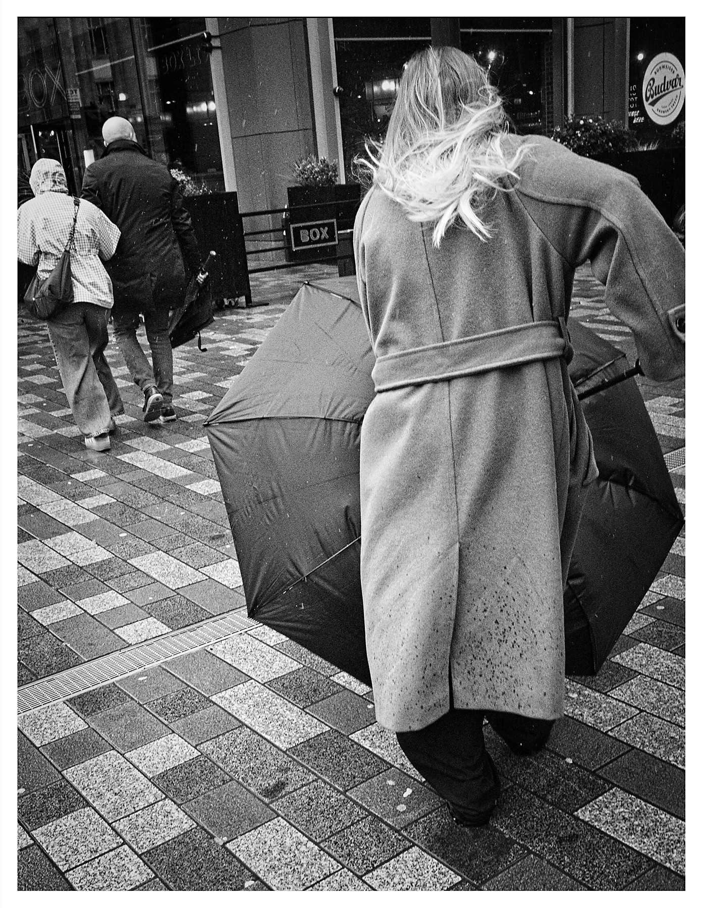 Black and white street photograph of a woman seen from behind in a long belted coat, her blonde hair blowing wildly in the wind as she struggles with an inside-out umbrella on a wet city centre pavement, with two other pedestrians and shopfronts visible behind her.