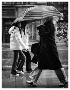 Black and white street photograph of two young women in white puffer jackets passing a blonde woman in a long dark coat on a rainy Manchester pavement, all carrying umbrellas, with a graffiti-tagged wall visible behind them.