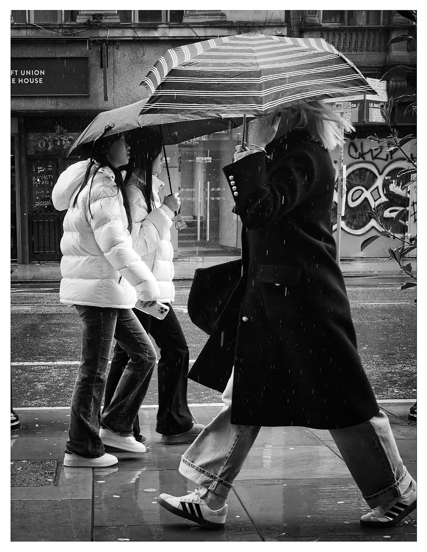 Black and white street photograph of two young women in white puffer jackets passing a blonde woman in a long dark coat on a rainy Manchester pavement, all carrying umbrellas, with a graffiti-tagged wall visible behind them.