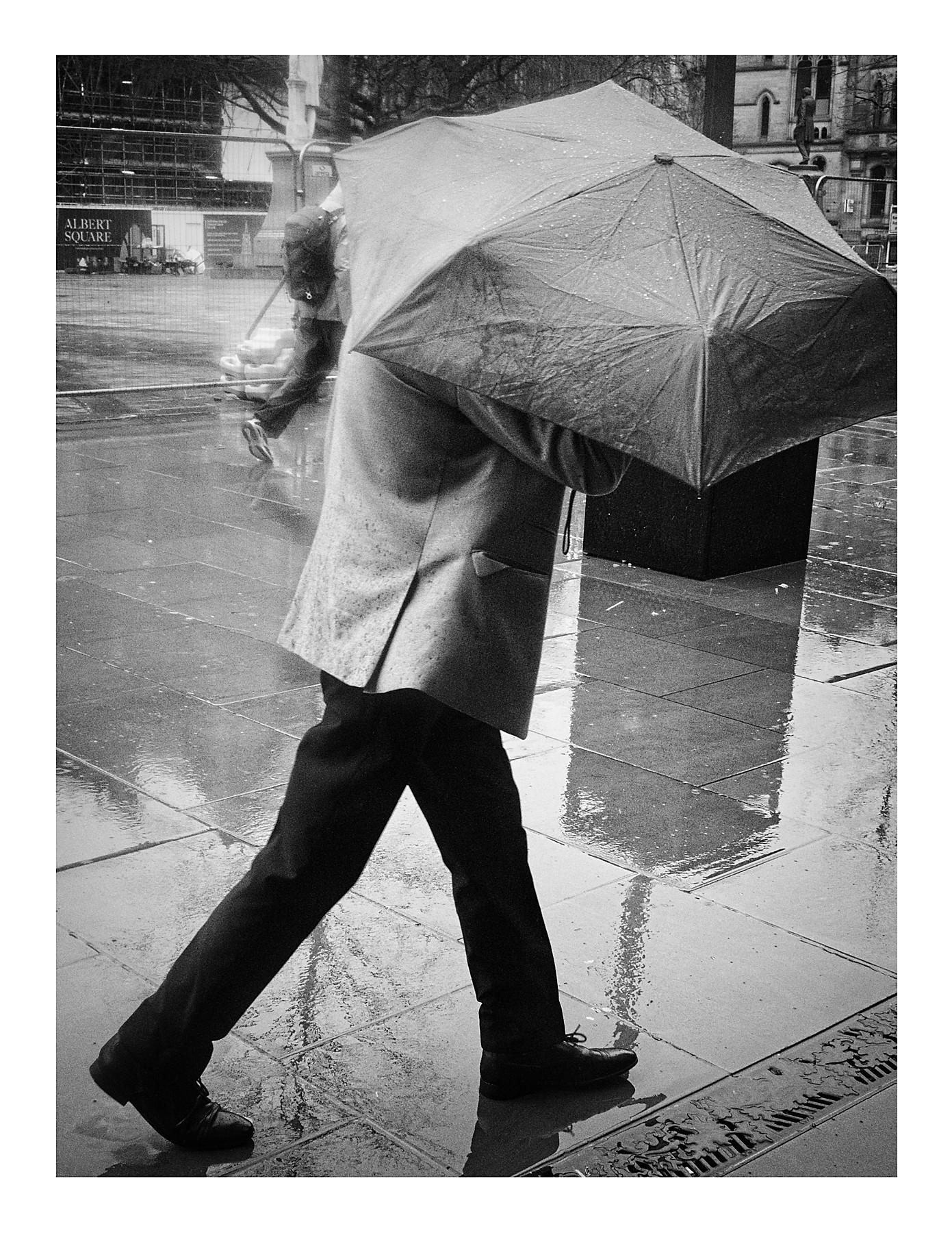 Black and white street photograph of a man walking in heavy rain in Albert Square, Manchester, his umbrella held so low it completely obscures his face and upper body, with his reflection visible in the wet paving slabs.